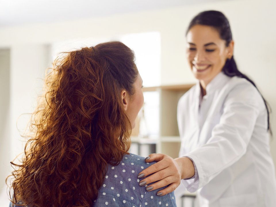 Stehende Ärztin in weissem Kittel legt mitfühlend die linke Hand auf die rechte Schulter einer vor ihr sitzenden Patientin. Der fröhliche Gesichtsausdruck der Ärztin vermittelt Zuversicht in guten Händen zu sein. 
