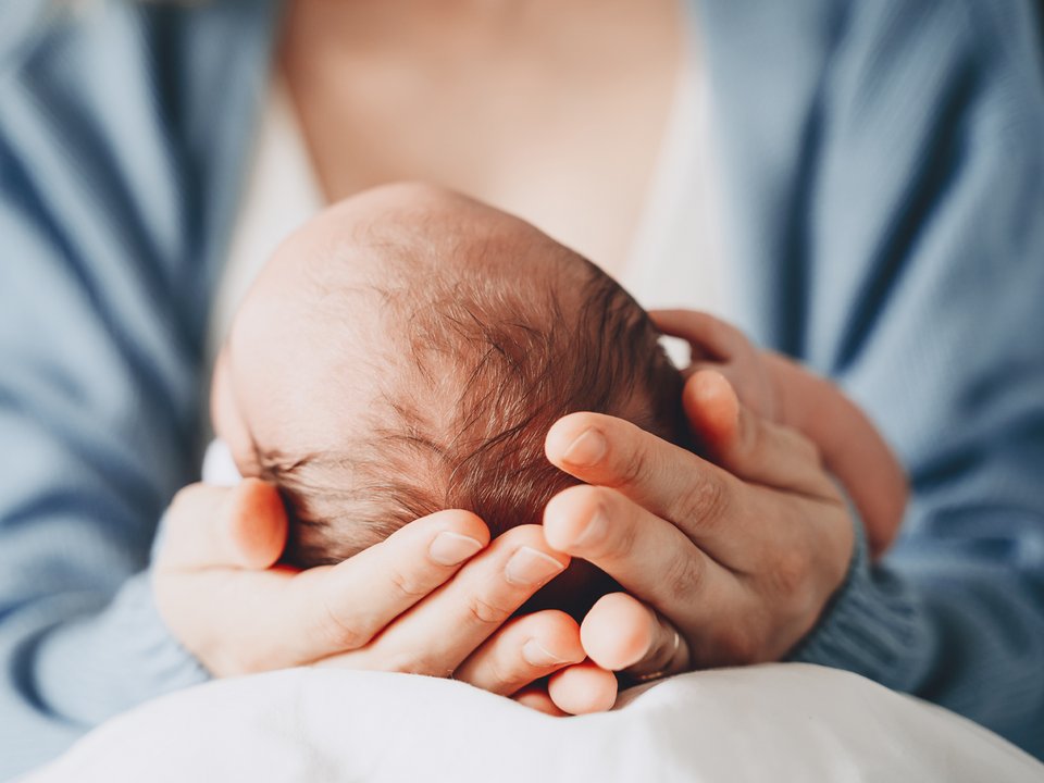 Frisch gewordene, sitzende Mutter hält ihr neugebohrenes Kind in beiden Händen auf ihrem Schoss. Symbolbild für die begleitende Schwangerschaft und Geburt in der Frauenklinik und soll Sicherheit und Geborgenheit ausstrahlen. 