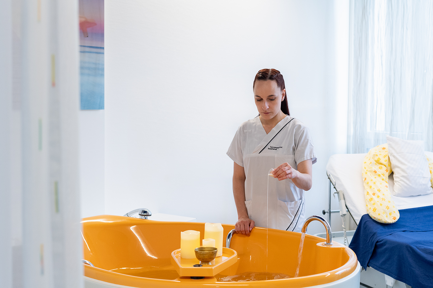 Hebamme lässt Öl ins Wasser ein und richtet alles für eine Wassergeburt ein. Auf der Geburtswanne stehen drei weisse Kerzen und eine Klangschale ünd verströhmt eine Wohlfühlatmosphäre 