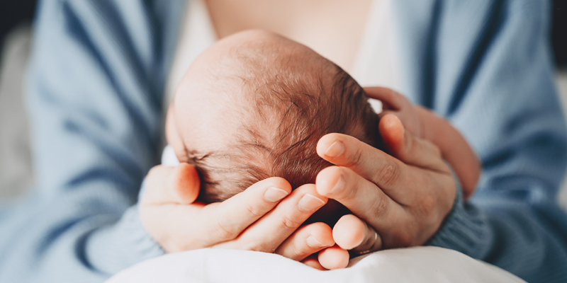 Frisch gewordene, sitzende Mutter hält ihr neugebohrenes Kind in beiden Händen auf ihrem Schoss. Symbolbild für die begleitende Schwangerschaft und Geburt in der Frauenklinik und soll Sicherheit und Geborgenheit ausstrahlen. 